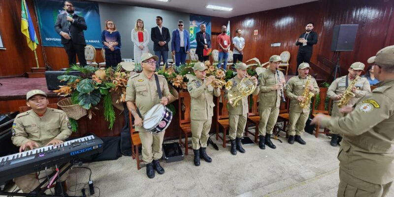 Foto horizontal da abertura da Quinta Conferência Estadual dos Direitos da Pessoa com Deficiência do Amapá. Dentro de um auditório com teto branco, paredes em madeira e piso cinza, à frente de um palco em madeira, em pé, uma banda composta por oito militares, tocam instrumentos de sopro, percussão e um teclado, sendo regidos por um maestro também militar. Todos estão vestidos com bonés e fardas marrons claro e coturnos pretos. Sobre o palco, em pé, oito pessoas que compuseram o painel de autoridades do evento. À direita o intérprete de libras usando roupa preta. Ao fundo e à esquerda, a bandeira do Estado do Amapá e um banner com o brasão do Governo do Estado.