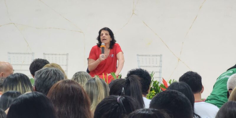 Foto horizontal, em ambiente interno, na palestra magna na V Conferência Estadual dos Direitos da Pessoa com Deficiência do Piauí. Com fundo branco e sobre um palco, falando ao microfone, a Secretária Nacional dos Direitos das Pessoas com Deficiência, Sra. Anna Paula Feminella. Ela é mulher, branca, cabelos pretos na altura dos ombros. Anna usa um vestido vermelho e um anel na mão direita, a mesma mão que segura o microfone. À sua frente, pessoas assistem à palestra.