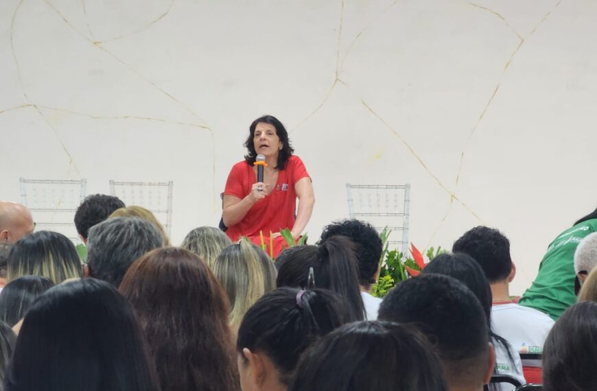 Foto horizontal, em ambiente interno, na palestra magna na V Conferência Estadual dos Direitos da Pessoa com Deficiência do Piauí. Com fundo branco e sobre um palco, falando ao microfone, a Secretária Nacional dos Direitos das Pessoas com Deficiência, Sra. Anna Paula Feminella. Ela é mulher, branca, cabelos pretos na altura dos ombros. Anna usa um vestido vermelho e um anel na mão direita, a mesma mão que segura o microfone. À sua frente, pessoas assistem à palestra.