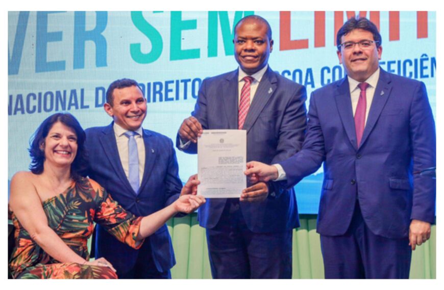 Foto horizontal, em ambiente interno, durante a V Conferência Estadual dos Direitos da Pessoa com Deficiência do Piauí e cerimônia de adesão do Novo Viver Limite no Estado do Piauí. Da esquerda para direita, a Secretária Nacional dos Direitos das Pessoas com Deficiência, Sra. Anna Paula Feminella. Ela é mulher, branca, cabelos pretos na altura dos ombros. Anna usa um vestido com uma estampa tropical, com imagens de folhas e está sentada em sua cadeira de rodas. O secretário para Inclusão da Pessoa com Deficiência do Estado do Piauí, Mauro Eduardo. Ele é um homem branco, cabelos castanhos e curtos, usa um terno xadrez azul, camisa branca e grava azul claro. O Ministro de Estado dos Direitos Humanos e da Cidadania, Sr. Silvio Luiz de Almeida. Ele é homem, negro, cabeça raspada, usa um terno cinza com uma camisa branca, gravata xadrez vermelha com branco e azul. O governador do Piauí, Rafael Fonteles, homem branco, cabelos castanhos e curtos, usa óculos de grau e terno azul, com camisa branca e gravata vinho. Todos estão sorrindo e ajudam o ministro a segurar o termo de adesão assinado pelas autoridades presentes. Ao fundo, o telão em led projeta a logomarca do Novo Viver sem Limite.