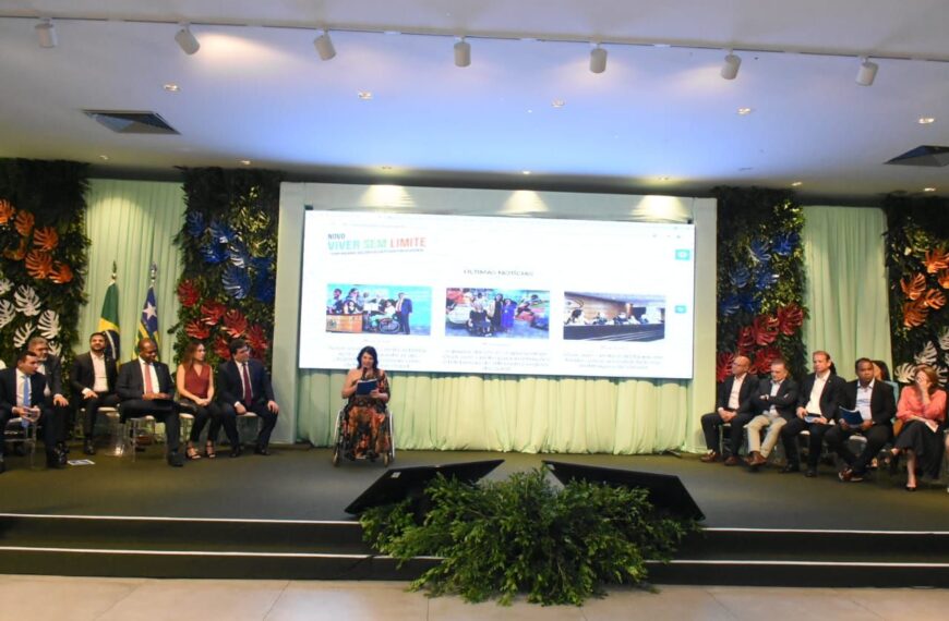 Foto horizontal, em ambiente interno. Evento de adesão do Novo Viver sem Limite no Estado do Piauí. Ao centro, a Secretária Nacional dos Direitos das Pessoas com Deficiência do Ministério dos Direitos Humanos e da Cidadania, Sra. Anna Paula Feminella. Ela é uma mulher, branca, cabelos pretos na altura dos ombros, usa um vestido com estampa tropical, com imagens de folhas nas cores laranja e verde. Anna está sentada em sua cadeira de rodas e, sobre um palco com piso preto e um arranjo de plantas ao centro, ela discursa segurando o microfone com a mão direita enquanto lê um documento que está na outra mão. À esquerda e à direita, sentadas, as pessoas que compuseram o painel de autoridades do evento. A parede de fundo é dividida por colunas de plantas alternando com cortina verde claro, onde à esquerda temos as bandeiras do Brasil e do Piauí e ao centro, um painel de LED onde está projetado a logo e imagens do Novo Viver sem Limites.