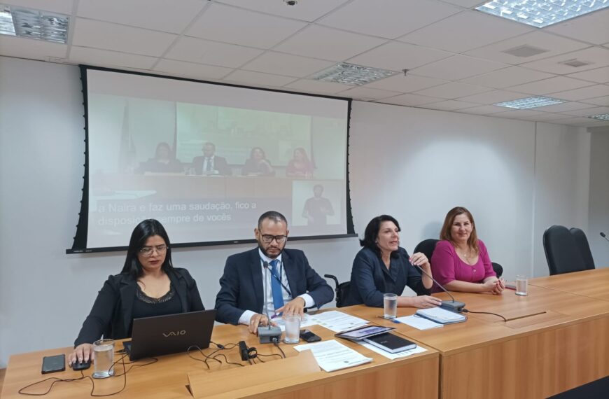 Foto horizontal em ambiente interno. Compondo o painel do grupo de trabalho da avaliação biopsicossocial quatro pessoas sentadas atrás de uma mesa em madeira com microfones. Da esquerda para direita, a assistente administrativa Tatiane Pereira de Araújo. Ela é mulher, parda, cabelos pretos e compridos, usa óculos de grau, blazer preto e blusa preta. O servidor Hisaac Alves de Oliveira. Ele é homem, pardo, cabelos e braba castanhos e curtos, usa óculos de grau, blazer azul escuro, camisa branca e gravata azul. A Secretária Nacional dos Direitos das Pessoas com Deficiência, Sra. Anna Paula Feminella. Ela é mulher, branca, cabelos pretos na altura dos ombros, usa blazer cinza escuro. A Diretora dos Direitos das pessoas com Deficiência, Sra. Naira Rodrigues Gaspar. Ela é mulher, branca, cabelos loiros e compridos, usa uma blusa rosa. A Secretária Anna Paula está falando ao microfone. Ao fundo, sobre a parede branca, uma tela de projetor com imagem da própria composição da mesa, legendas e um intérprete de libras.