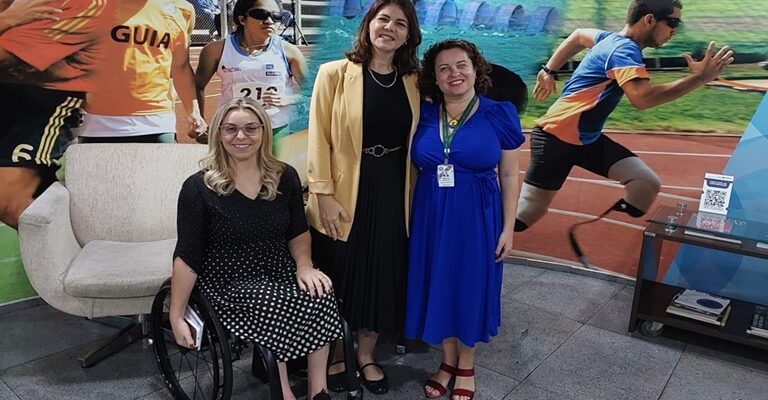 Foto horizontal, em ambiente interno, da comitiva que acompanhou a Conferência Estadual da Pessoa com Deficiência de São Paulo. No centro da imagem, três pessoas. À esquerda, Izana Figueira, mulher, branca, cabelos lisos, loiros e compridos. Ela usa óculos de grau com armação bege e está sorrindo. Izana está com vestido preto com bolinhas brancas, tênis preto e branco e está em sua cadeira de rodas preta. No meio, em pé, Maria Aparecida Correia, mulher, branca, cabelos lisos, castanhos, na altura dos ombros e está sorrindo. Ela usa blazer amarelo claro sobre um vestido preto comprido, cinto de corrente prata e sapatos pretos. Maria Aparecida está usando uma corrente prata no pescoço. À direita, em pé, Daiane Mantoanelli, mulher, branca, cabelos castanhos, cacheados, na altura dos ombros e está sorrindo. Ela usa um vestido comprido azul, sandálias com tiras vermelhas. No pescoço, Daiane usa seu crachá com fita verde e um colar com um ipê amarelo no pingente. Atrás da Izana, uma poltrona bege. Ao fundo, um banner grande que vai de lado a lado da foto com imagens de atletas paralímpicos em competições, e o piso da sala é cinza.