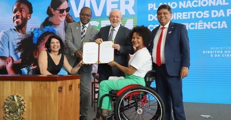 Foto horizontal, em ambiente interno, do evento de lançamento do Novo Viver sem Limite, com as pessoas que compuseram o palco. Da esquerda para direita, atrás de um púlpito em madeira com brasão da república dourado, a Secretária Nacional dos Direitos das Pessoas com Deficiência, Sra. Anna Paula Feminella. Ela é mulher, branca, cabelos pretos na altura dos ombros. Anna usa uma blusa sem mangas preta e brincos prateados. Ela sorri e está em sua cadeira de rodas. Em pé, o Ministro de Estado dos Direitos Humanos e da Cidadania, Sr. Silvio Luiz de Almeida. Ele é homem, negro, cabeça raspada, usa óculos de grau e está sorrindo. Silvio está com terno cinza claro, camisa branca, gravata xadrez cinza com branco e sapatos pretos. Em pé, o Presidente da República, Sr. Luiz Inácio Lula da Silva. Ele é homem, branco, cabelos e barba grisalhos. Lula usa terno cinza escuro, camisa branca e gravata listrada preto e cinza. Sra. Agna Alves da Cruz, mulher, negra, cabelos pretos, crespos e curtos e está sorrindo. Ela usa uma blusa branca de mangas curtas, calça na cor verde claro e está em sua cadeira de rodas preta e vermelha. Em pé, o Deputado Federal, Sr. Márcio Jerry. Ele é homem, pardo, cabelos castanhos, curtos e está sorrindo. Márcio usa um terno azul escuro, camisa branca, gravata vermelha e sapatos marrons. Todas as pessoas seguram um documento de duas folhas aberto como se fosse um livro. O fundo é um banner azul claro que vai de lado a lado da foto, com três pessoas e está escrito Novo Viver sem Limite, Plano Nacional dos Direitos das Pessoas com Deficiência, Ministério dos Direitos Humanos e da Cidadania. O piso do palco é de carpete cinza.