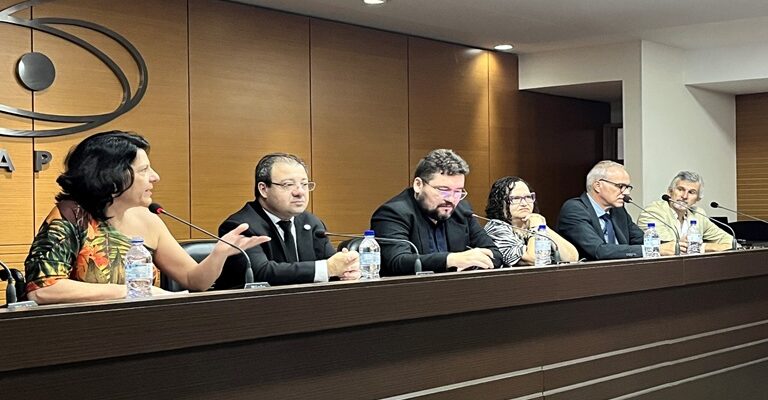 Foto horizontal, em ambiente interno, no Fórum sobre Acessibilidade nas Instituições Públicas de Educação Superior e Profissional Tecnológica. Na composição da mesa, todas as pessoas estão sentadas atrás de uma bancada em madeira com três listas cinzas na horizontal, tem um microfone e uma garrafa com água à frente de cada integrante. Da esquerda para direita, a Secretária Nacional dos Direitos das Pessoas com Deficiência do Ministério dos Direitos Humanos e da Cidadania, Sra. Anna Paula Feminella. Ela é uma mulher, branca, cabelos pretos na altura dos ombros. Anna usa uma blusa estampada com folhas de plantas, nas cores laranjada e verde, com manga curta no lado direito e sem manga no lado esquerdo. Ela está falando ao microfone. O Diretor de Políticas e Programas de Educação Superior substituto, da Secretaria de Educação Superior do ministério da Educação, Ricardo Portocarrero. Ele um é homem, branco, cabelos grisalhos e curtos, usa óculos de grau, terno preto, camisa branca e gravata preta. O Diretor de Políticas de Educação Especial na Perspectiva Inclusiva do ministério da Educação, Sr. Alexandre Mapurunga. Ele é um homem, branco, cabelos e barba pretos e curtos, usa óculos de grau, terno preto e camisa azul escuro. A Diretora de Formação de Professores de Educação Básica da Coordenação de Aperfeiçoamento de Pessoal de Nível Superior do Ministério da Educação, Sra. Marcia Ferreira. Ela é uma mulher, branca, cabelos cacheados e pretos na altura dos ombros, usa uma blusa branca com listras pretas. O Reitor da Universidade Federal do Rio Grande do Norte, José Daniel Diniz Melo. Ele é um homem branco, cabelos grisalhos e curtos, usa óculos de grau com armação preta, terno azul escuro, camisa azul claro e gravata azul escuro. O coordenador do evento, Francisco Ricardo Lins Vieira de Melo, da UFRN. Ele é um homem branco, cabelos e cavanhaque grisalhos, usa um blazer bege e camisa branca.