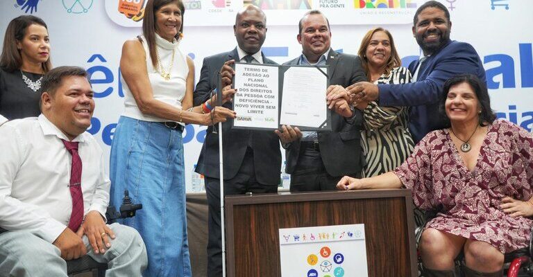 Na foto, há um grupo de oito pessoas sorridentes em um evento. No centro, um homem de terno segura um documento com as palavras "TERMO DE ADESÃO AO PLANO NACIONAL DOS DIREITOS DA PESSOA COM DEFICIÊNCIA - VIVER SEM LIMITE". À esquerda, um homem sentado em uma cadeira de rodas e uma mulher de pé, vestindo uma blusa branca e saia jeans, sorriem enquanto olham para o documento. À direita, um homem de camisa azul e uma mulher de vestido preto e branco também sorriem. No canto inferior direito, uma mulher sentada em uma cadeira de rodas sorri para a câmera. No fundo, há banners com logotipos do governo e símbolos de acessibilidade. No canto inferior esquerdo, há um banner com o texto "V Conferência Estadual" e ícones coloridos representando diferentes tipos de deficiência.