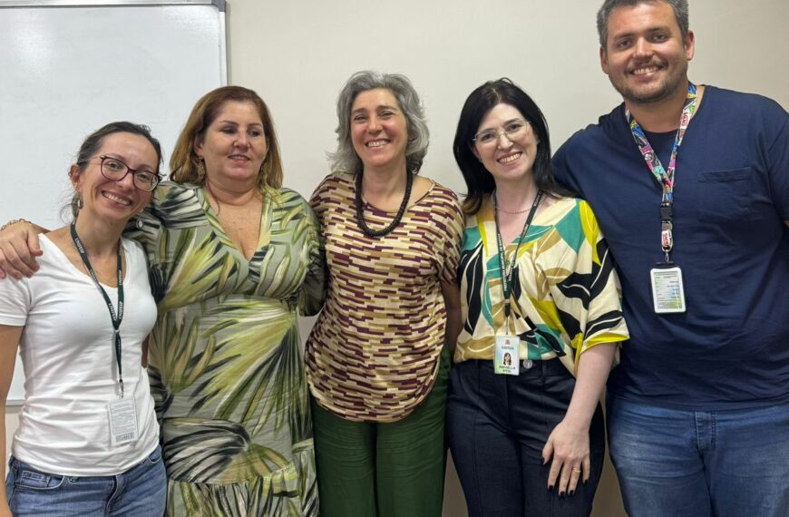 Na foto, há cinco pessoas sorrindo para a câmera, quatro mulheres e um homem, em pé em frente a uma lousa branca em uma sala com paredes claras. Da esquerda para a direita: a primeira mulher está usando uma camiseta branca e jeans, com um crachá pendurado no pescoço; a segunda mulher tem cabelos loiros, usa um vestido com estampa verde e bege e também tem um crachá, já a terceira mulher tem cabelos grisalhos, usa uma blusa estampada em tons de marrom e calças verdes, com um crachá. Em seguida, uma quarta mulher usa óculos, uma blusa com estampa geométrica colorida, calças pretas e um crachá, enquanto o homem, à direita, tem cabelos castanhos curtos, usa uma camiseta azul, jeans e tênis vermelhos, e também tem um crachá. Todos são representantes dos movimentos das pessoas com deficiência e parecem estar em um ambiente de trabalho e estão usando crachás de identificação.