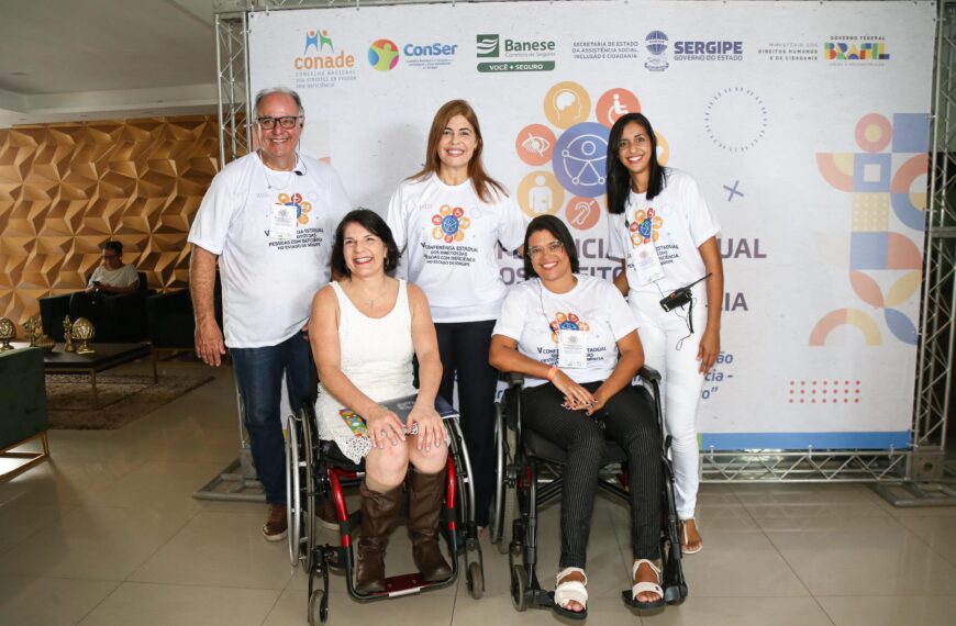 Na foto, há cinco pessoas sorridentes posando para uma foto em um evento. Duas mulheres estão sentadas em cadeiras de rodas na frente, ambas vestindo roupas brancas; a mulher à esquerda, que é a secretária nacional dos Direitos da Pessoa com Deficiência, Anna Paula Feminella, usa um vestido branco e botas marrons, enquanto a mulher à direita usa uma camiseta branca e calças pretas. Atrás delas, há três pessoas em pé, um homem e duas mulheres, todos vestindo camisetas brancas com a mesma estampa. O fundo é um painel com logotipos de patrocinadores e o texto "V CONFERÊNCIA ESTADUAL DOS DIREITOS DA PESSOA COM DEFICIÊNCIA". Há um sofá e uma mesa de centro à esquerda no fundo, e o ambiente parece ser um espaço de conferência ou evento