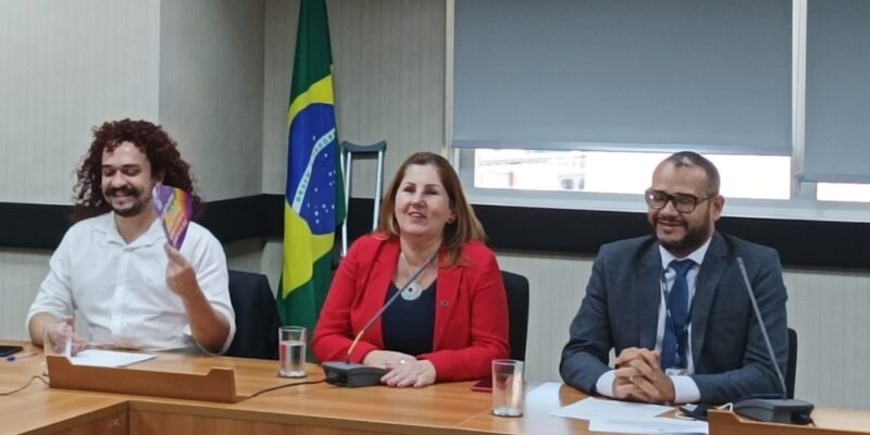 Na imagem, três pessoas estão sentadas em uma mesa de reunião em uma sala com uma bandeira do Brasil ao fundo. À esquerda, um homem com cabelos cacheados e barba, vestindo uma camisa branca, sorri enquanto segura um leque colorido. No centro, uma mulher com cabelos loiros e vestindo um blazer vermelho, também sorri. À direita, um homem com cabelos curtos e barba, vestindo um terno azul escuro e gravata, sorri enquanto olha para o lado. Todos parecem estar em um ambiente formal e descontraído.