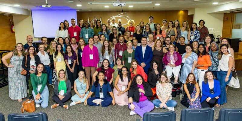 A imagem mostra um grande grupo de pessoas posando para uma foto em um ambiente interno. Há aproximadamente 50 pessoas, homens e mulheres, de diversas idades e etnias. Eles estão em pé e sentados em várias fileiras, sorrindo para a câmera. Alguns usam crachás pendurados no pescoço. Ao fundo, há uma tela de projeção branca e um projetor no teto. O local parece ser uma sala de conferências ou um auditório, com cadeiras pretas empilhadas à frente e carpete no chão.