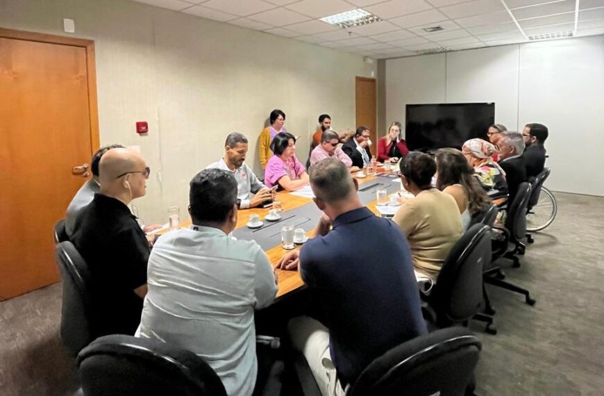 A imagem mostra uma sala de reuniões com várias pessoas sentadas ao redor de uma grande mesa retangular. Todos os integrantes da mesa integram o Conselho Nacional dos Direitos da Pessoa com Deficiência (Conade). A sala tem paredes claras e um teto com várias luminárias embutidas. As pessoas parecem estar envolvidas em uma discussão ou apresentação, algumas delas com copos de água à frente. No canto direito, há uma pessoa em uma cadeira de rodas. Trata-se da presidente do Conade e secretária nacional dos Direitos da Pessoa com Deficiência, Anna Paula Feminella. Ao fundo, pode-se ver uma porta de madeira fechada e uma tela de televisão desligada pendurada na parede.