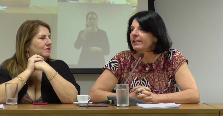 A imagem mostra duas mulheres sentadas lado a lado em uma mesa, participando de uma reunião ou conferência. A mulher à esquerda tem cabelo castanho claro e está usando uma blusa preta com um decote em V. Ela está com as mãos cruzadas e parece estar ouvindo atentamente. A mulher à direita tem cabelo preto e está usando uma blusa estampada em tons de vermelho e marrom. Ela está falando ao microfone e tem alguns papéis à sua frente. Na mesa, há copos de água e uma xícara de café. Ao fundo, há uma tela de projeção com a imagem de uma intérprete de Libras (Língua Brasileira de Sinais)