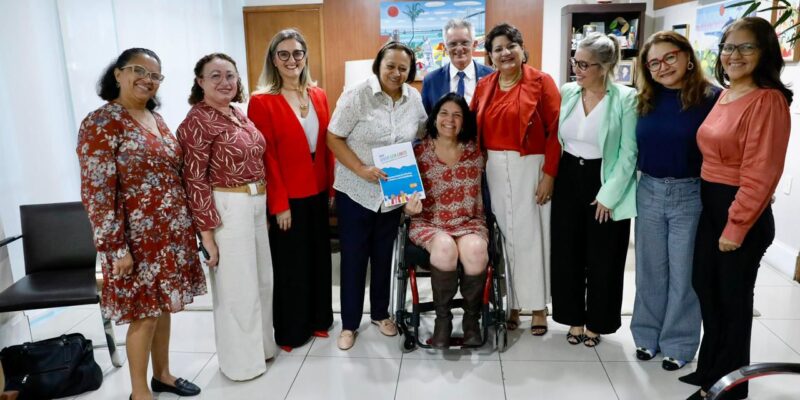 Na foto, há um grupo de nove pessoas em um ambiente interno, possivelmente um escritório ou sala de reuniões. Quatro homens e cinco mulheres estão sorrindo para a câmera. Uma das mulheres está sentada em uma cadeira de rodas no centro do grupo e segura um livro com a capa azul que diz "Educação Inclusiva". Todos estão bem vestidos, sugerindo um evento formal ou uma reunião profissional. O ambiente tem decoração simples, com uma grande janela ao fundo coberta por cortinas brancas e alguns quadros na parede.