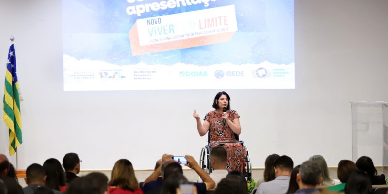 A imagem mostra um seminário com uma palestrante mulher sentada em uma cadeira de rodas. Ela está no centro, em frente a uma grande tela que exibe a frase "Seminário de apresentação" e abaixo "Novo Viver Sem Limite - Plano Nacional dos Direitos da Pessoa com Deficiência". A tela também mostra logos de patrocinadores e a bandeira do Brasil ao lado esquerdo. A palestrante, que tem cabelos pretos e usa um vestido estampado, gesticula enquanto fala para uma plateia atenta, composta por diversas pessoas sentadas olhando para ela.