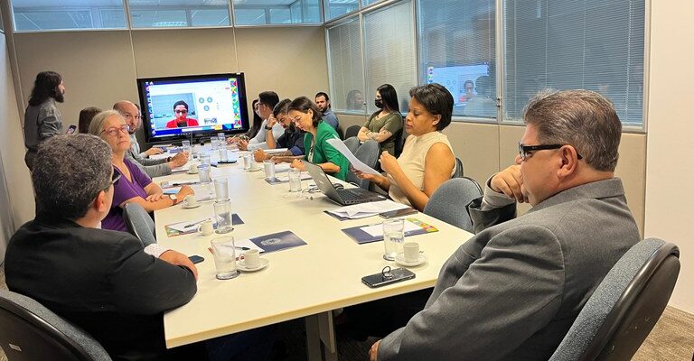 A imagem mostra uma sala de reuniões com várias pessoas sentadas ao redor de uma mesa retangular. A mesa está coberta com copos de água, xícaras de café, laptops e documentos. Há um monitor grande na parede ao fundo, exibindo uma videoconferência com a imagem de um homem de camiseta vermelha. As pessoas na sala parecem estar prestando atenção na tela ou em documentos. A sala tem paredes de vidro e iluminação fluorescente.