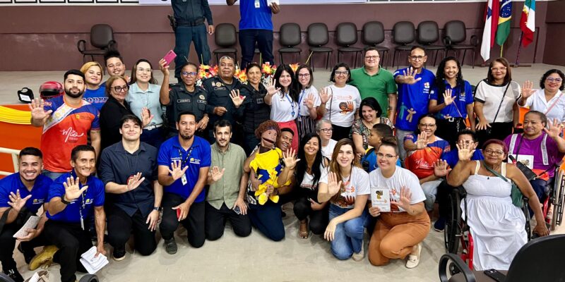 A imagem mostra um grupo grande de pessoas posando para uma foto em um ambiente interno, possivelmente um auditório ou sala de conferências. Há cerca de 30 pessoas na foto, incluindo homens e mulheres de diferentes idades. Algumas pessoas estão em pé, enquanto outras estão agachadas ou sentadas na frente. No fundo, há uma tela de projeção e várias cadeiras vazias. Algumas pessoas estão vestindo uniformes e outras estão usando camisetas de diferentes cores, algumas com logotipos ou mensagens. Muitas pessoas estão sorrindo e fazendo o gesto de "cinco" com a mão levantada, como se estivessem participando de uma campanha ou evento específico. Há também bandeiras do Brasil e de outros estados ou instituições no lado direito da imagem. A atmosfera parece ser de celebração ou conscientização, com todos demonstrando entusiasmo e união.