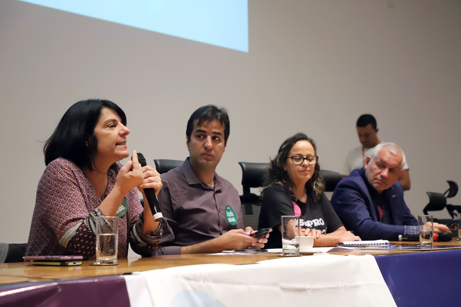 A imagem mostra quatro pessoas sentadas lado a lado em uma mesa de conferência. Da esquerda para a direita: 1. Uma mulher de cabelo preto e curto, usando uma blusa estampada, está falando ao microfone. Na frente dela, há um copo de água e um celular sobre a mesa; 2. Um homem de cabelo preto e curto, vestindo uma camisa roxa, está olhando para a mulher que fala. Ele tem um adesivo verde no peito; 3. Uma mulher de cabelo castanho e ondulado, usando óculos e uma camiseta preta com algo escrito, está olhando para a frente; e 4. Um homem de cabelo grisalho, vestindo um terno azul, está olhando para a mulher ao seu lado. Na frente dele, há um copo de água e um caderno. Ao fundo, há uma pessoa em pé, desfocada, que parece estar mexendo no celular. A parede de fundo é branca.