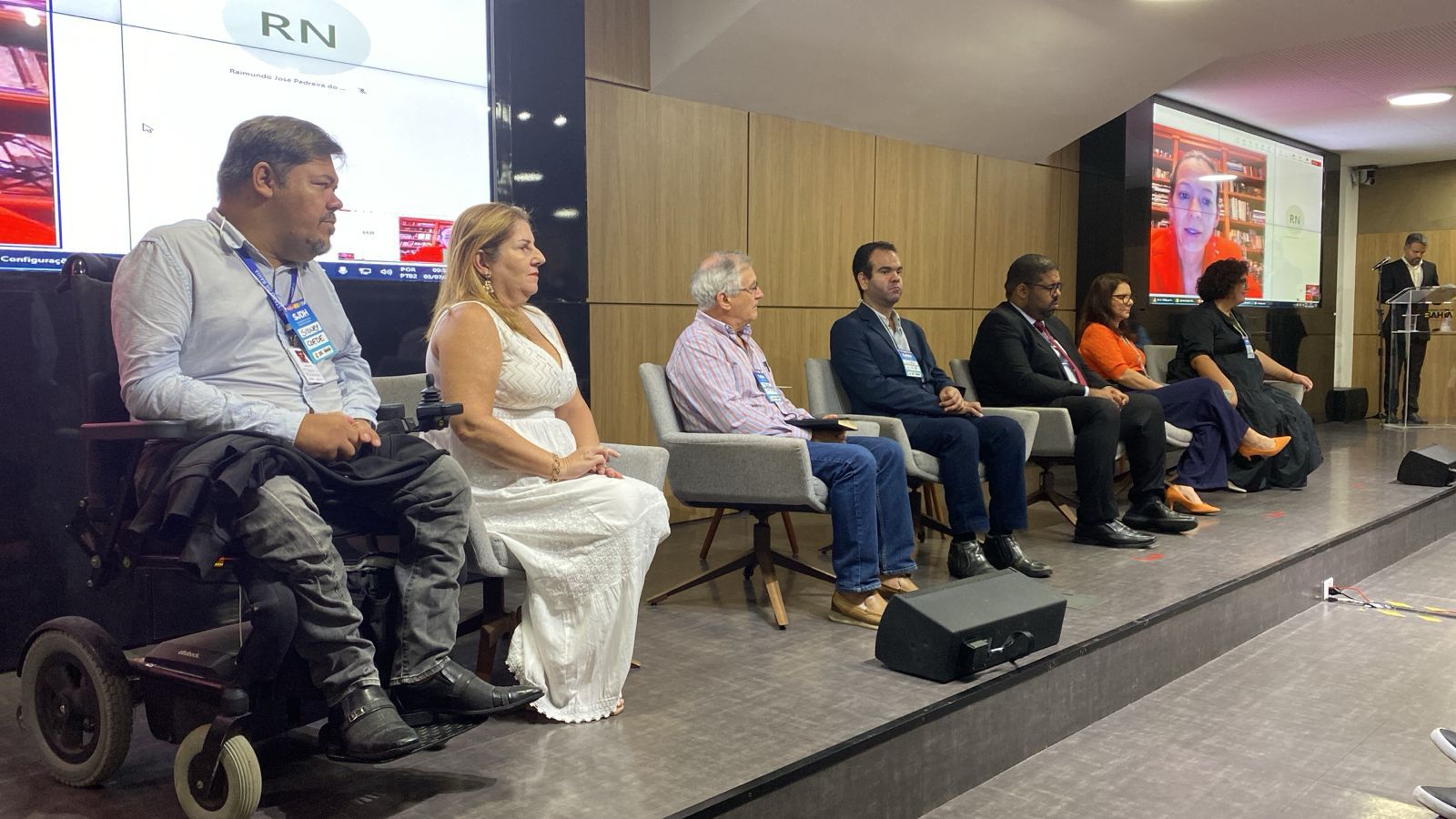 A imagem mostra um painel de discussão com sete pessoas sentadas em cadeiras em um palco. Da esquerda para a direita: um homem em uma cadeira de rodas elétrica, vestindo uma camisa social clara e calça escura; uma mulher de vestido branco, com cabelo loiro; um homem idoso de camisa listrada rosa e calça jeans; um homem de terno escuro e camisa clara; um homem de terno preto e gravata vermelha; uma mulher de blusa laranja e calça escura; uma mulher de vestido preto e sapatos laranja. Ao fundo, há uma tela grande exibindo uma videoconferência com a imagem de uma mulher de blusa vermelha. À direita do palco, um homem de terno está em pé, falando em um púlpito. O ambiente parece ser uma sala de conferências com paredes de madeira clara.