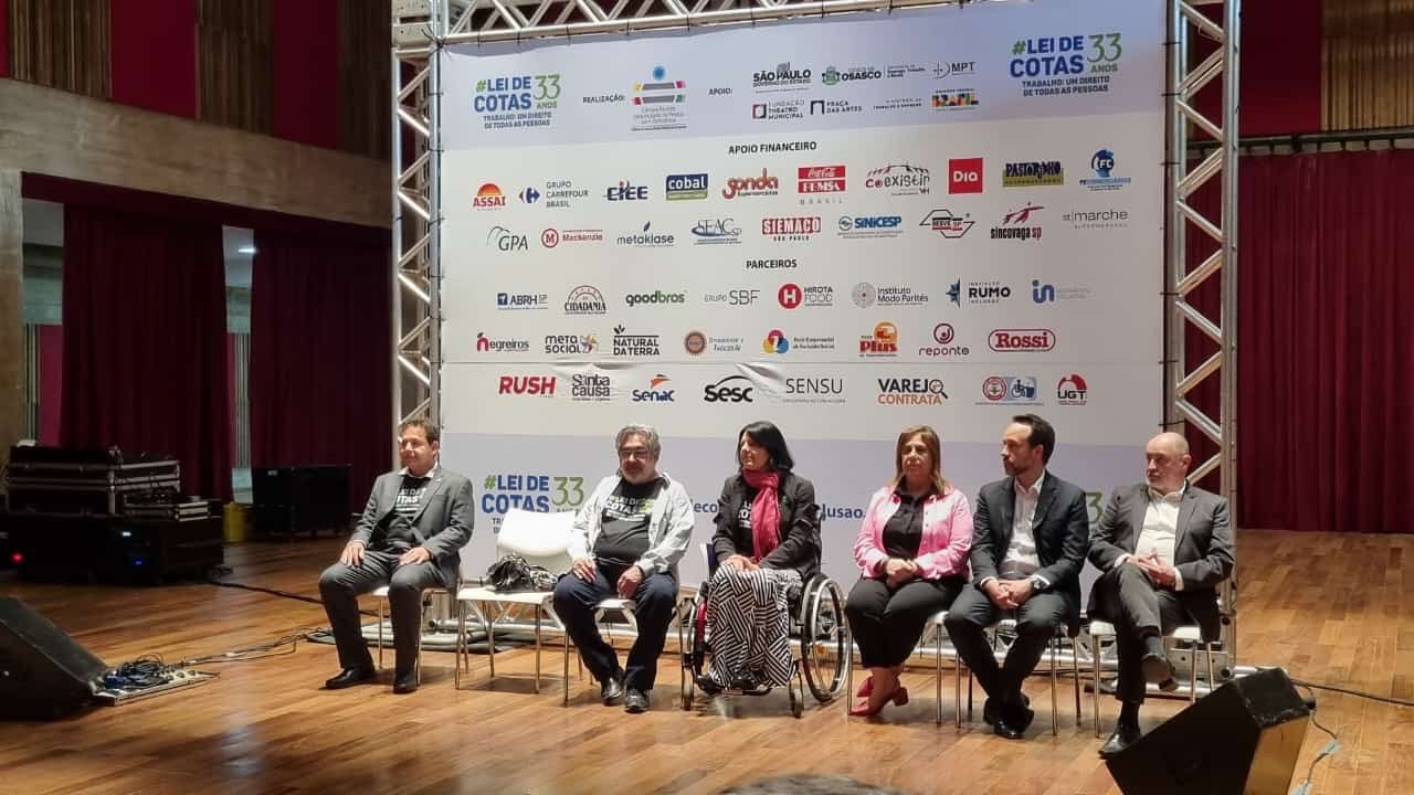 A imagem mostra um evento em um auditório. No palco, há seis pessoas sentadas em cadeiras, lado a lado. Atrás delas, há um grande painel branco com diversos logotipos de empresas e organizações. O painel também tem o texto "Lei de Cotas 33 anos" em destaque. As pessoas no palco estão vestidas formalmente, e uma delas está em uma cadeira de rodas. O ambiente parece ser um auditório com cortinas vermelhas ao fundo e piso de madeira. Há também equipamentos de som e iluminação no palco.