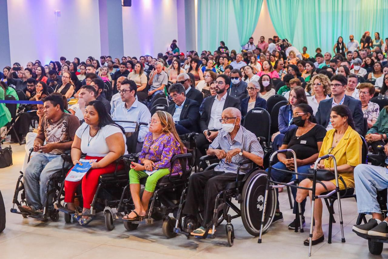 A imagem mostra uma grande audiência em um evento ou conferência. A sala está cheia de pessoas sentadas em cadeiras, com algumas pessoas em cadeiras de rodas na frente. A maioria das pessoas está olhando para a frente, possivelmente assistindo a uma apresentação ou discurso. As cortinas ao fundo são de cor verde clara, e há uma iluminação suave na sala. Algumas pessoas estão usando máscaras faciais. A diversidade de idades e estilos de roupas sugere que o evento é inclusivo e aberto a todos.