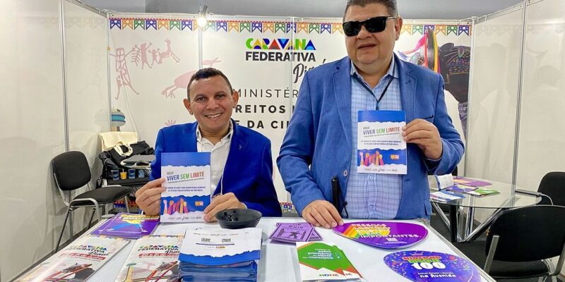 A imagem mostra duas pessoas em pé atrás de uma mesa em um estande de exposição. Ambos estão vestindo ternos azuis. A pessoa à esquerda está sorrindo e segurando um folheto com o título "Viver Sem Limite". A pessoa à direita está usando óculos escuros e também segurando um folheto semelhante. A mesa está coberta com vários materiais impressos, incluindo folhetos e panfletos. No fundo, há um banner com o logotipo do Governo Federal do Brasil e o texto "Ministério dos Direitos Humanos e da Cidadania". A decoração do estande inclui bandeirinhas coloridas no topo.