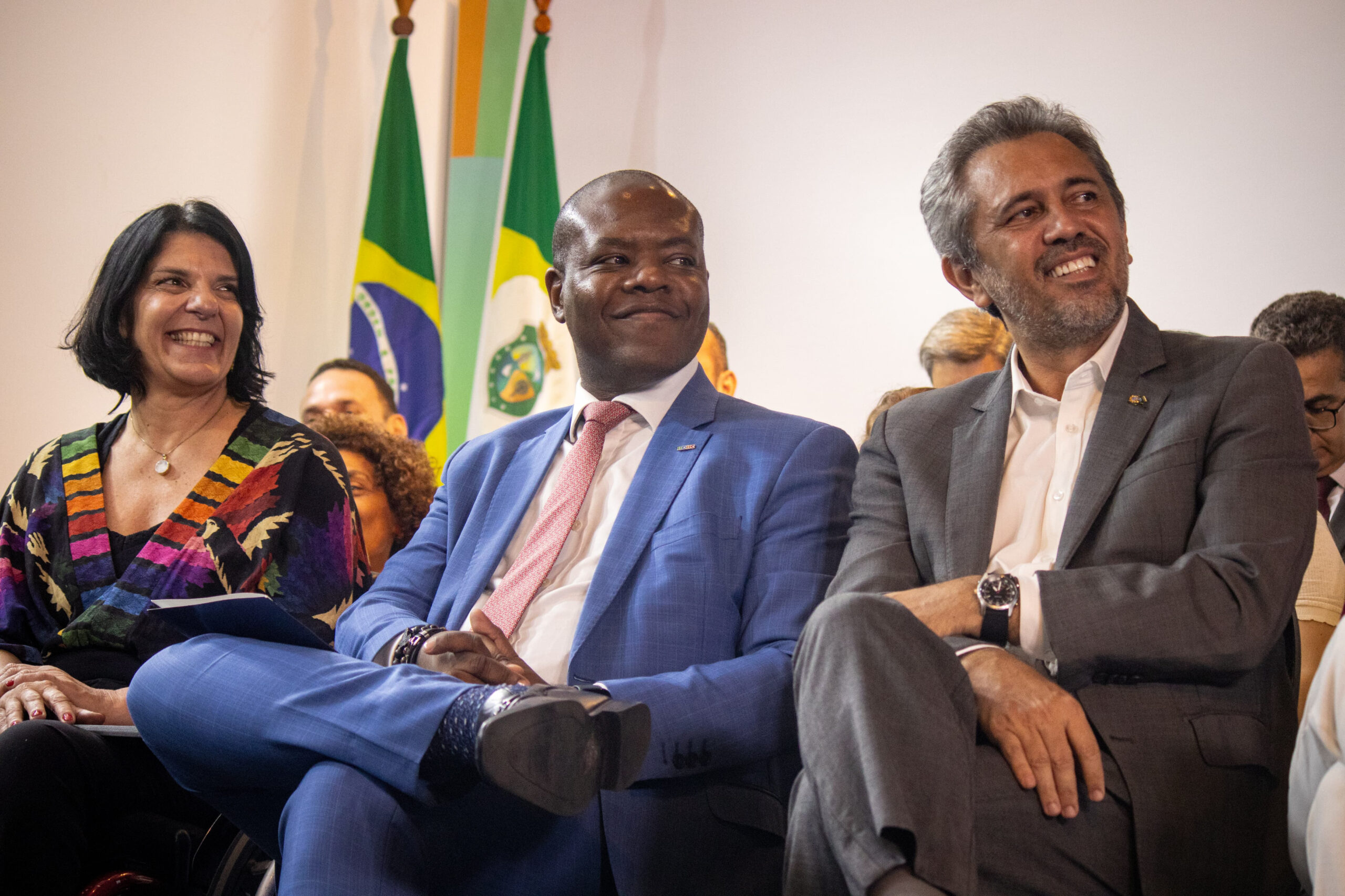 A imagem mostra três pessoas sentadas lado a lado, sorrindo. À esquerda, há uma mulher de cabelo preto e curto, vestindo uma blusa colorida com padrões geométricos. No centro, há um homem negro usando um terno azul claro e gravata rosa. À direita, há um homem de pele clara com cabelo grisalho, vestindo um terno cinza e camisa branca. Ao fundo, há bandeiras do Brasil e de um estado brasileiro. Todos parecem estar em um evento formal ou cerimônia.