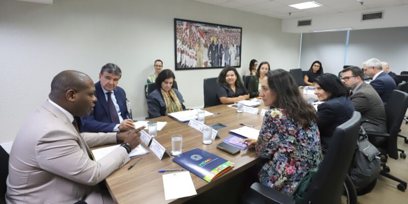 A imagem mostra uma reunião em uma sala de conferências. Há onze pessoas sentadas ao redor de uma mesa retangular de madeira. A maioria das pessoas está vestida formalmente, com ternos e roupas de trabalho. À esquerda, um homem negro de terno bege está falando. Ao lado dele, há um homem de terno azul escuro e gravata, e uma mulher de cabelo escuro e cachecol colorido. Do outro lado da mesa, há várias pessoas, incluindo uma mulher de blusa floral, uma mulher de cabelo curto e preto, e um homem de óculos e barba. No fundo da sala, há uma grande foto na parede que mostra uma cerimônia com várias pessoas vestidas de branco. A sala tem iluminação fluorescente e janelas com persianas fechadas. Na mesa, há papéis, pastas, copos de água e alguns dispositivos eletrônicos.