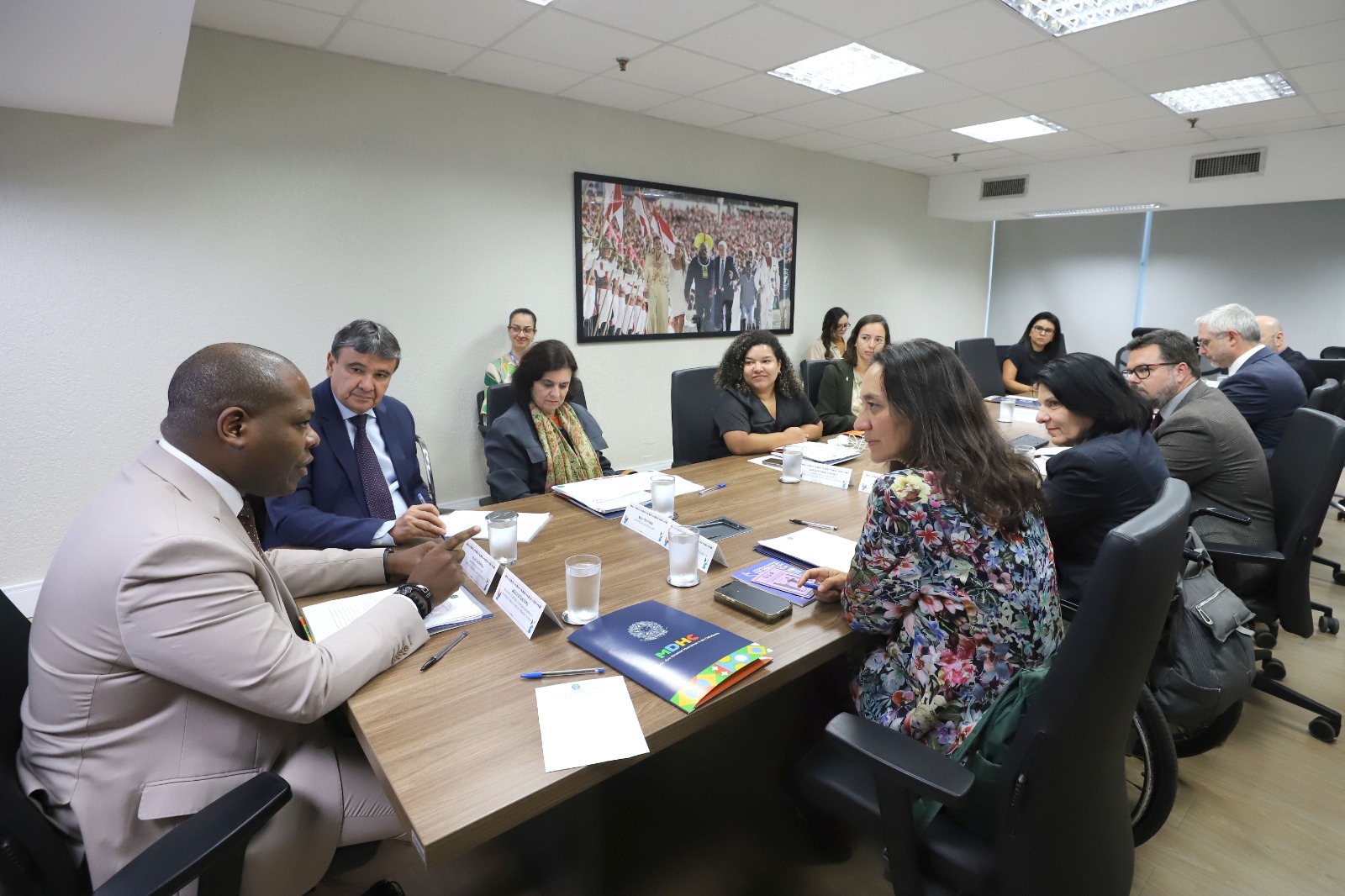 A imagem mostra uma reunião em uma sala de conferências. Há onze pessoas sentadas ao redor de uma mesa retangular de madeira. A maioria das pessoas está vestida formalmente, com ternos e roupas de trabalho. À esquerda, um homem negro de terno bege está falando. Ao lado dele, há um homem de terno azul escuro e gravata, e uma mulher de cabelo escuro e cachecol colorido. Do outro lado da mesa, há várias pessoas, incluindo uma mulher de blusa floral, uma mulher de cabelo curto e preto, e um homem de óculos e barba. No fundo da sala, há uma grande foto na parede que mostra uma cerimônia com várias pessoas vestidas de branco. A sala tem iluminação fluorescente e janelas com persianas fechadas. Na mesa, há papéis, pastas, copos de água e alguns dispositivos eletrônicos.