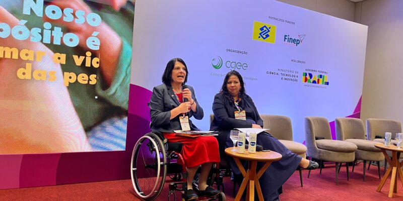 A imagem mostra duas mulheres sentadas em um palco durante um evento. A mulher à esquerda está em uma cadeira de rodas, vestindo um blazer azul escuro e uma saia vermelha. Ela está falando ao microfone. A mulher à direita está sentada em uma cadeira, vestindo um conjunto azul escuro, e está olhando para a outra mulher. Atrás delas, há um painel branco com vários logotipos, incluindo o da Finep, CGEE (Centro de Gestão e Estudos Estratégicos), Ministério da Ciência, Tecnologia e Inovação, e o Governo Federal do Brasil. À esquerda, há uma tela grande com a frase "Nosso propósito é transformar a vida das pessoas". No palco, há mesas pequenas com garrafas de água e copos. O chão do palco é coberto por um carpete vermelho.