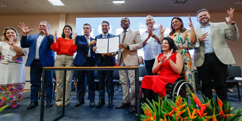 A imagem mostra um grupo de pessoas em um evento formal, posando para uma foto. No centro, dois homens estão segurando um documento aberto, que parece ser um contrato ou acordo. Eles estão sorrindo e vestem ternos. Ao redor deles, há outras pessoas, algumas aplaudindo e outras fazendo gestos com as mãos. À direita, uma mulher em uma cadeira de rodas está vestida com um vestido vermelho e também está aplaudindo. O ambiente parece ser um auditório ou sala de conferências, com um grande painel ao fundo e arranjos de flores tropicais na frente. Todos parecem estar celebrando ou comemorando algo importante.