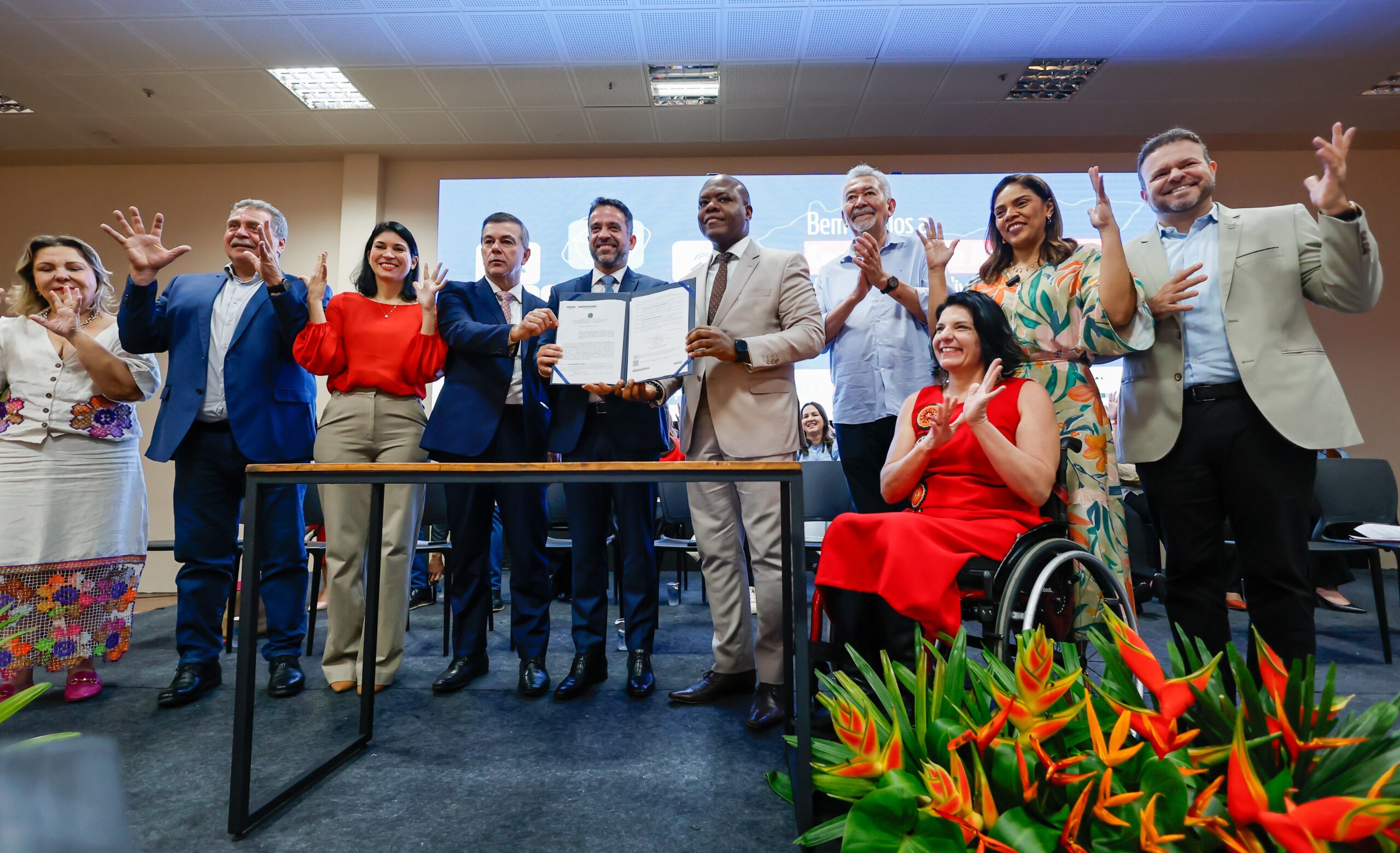 A imagem mostra um grupo de pessoas em um evento formal, posando para uma foto. No centro, dois homens estão segurando um documento aberto, que parece ser um contrato ou acordo. Eles estão sorrindo e vestem ternos. Ao redor deles, há outras pessoas, algumas aplaudindo e outras fazendo gestos com as mãos. À direita, uma mulher em uma cadeira de rodas está vestida com um vestido vermelho e também está aplaudindo. O ambiente parece ser um auditório ou sala de conferências, com um grande painel ao fundo e arranjos de flores tropicais na frente. Todos parecem estar celebrando ou comemorando algo importante.