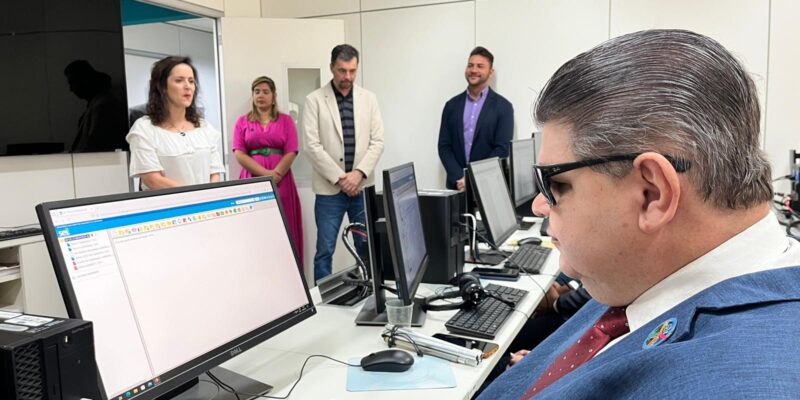 A imagem mostra um ambiente com várias pessoas. Em primeiro plano, à direita, há um homem de óculos escuros, cabelo grisalho penteado para trás, vestindo um terno azul e gravata vermelha com bolinhas brancas. Ele está sentado em frente a um computador, olhando para a tela. No fundo, há quatro pessoas em pé, duas mulheres e dois homens. A mulher à esquerda está vestindo uma blusa branca, a mulher ao lado dela está de vestido rosa, o homem ao lado dela veste um blazer bege e o último homem, à direita, veste um blazer azul escuro. Eles parecem estar observando ou participando de uma atividade no escritório. A sala tem paredes brancas e há um grande monitor de televisão na parede à esquerda.