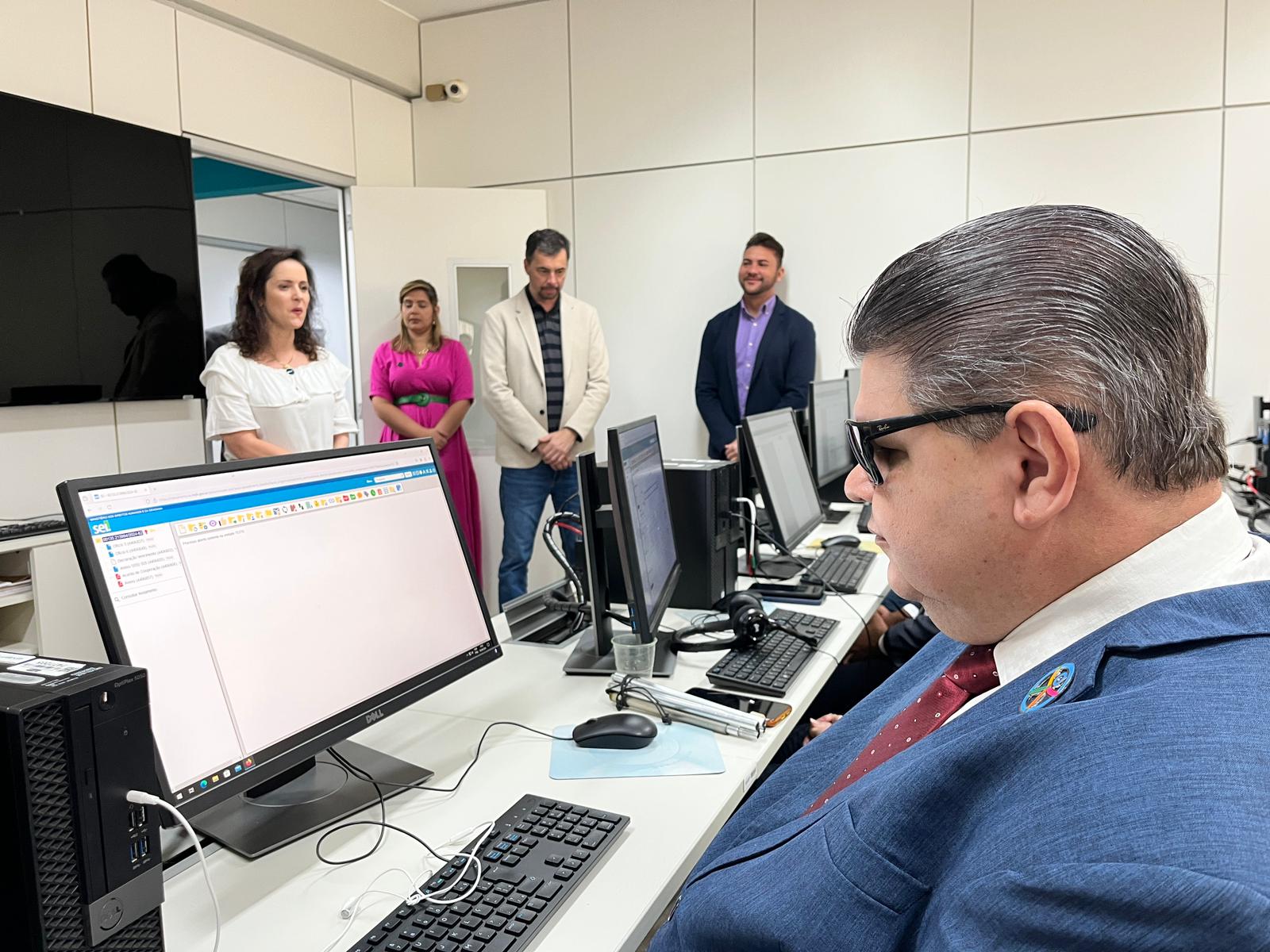 A imagem mostra um ambiente com várias pessoas. Em primeiro plano, à direita, há um homem de óculos escuros, cabelo grisalho penteado para trás, vestindo um terno azul e gravata vermelha com bolinhas brancas. Ele está sentado em frente a um computador, olhando para a tela. No fundo, há quatro pessoas em pé, duas mulheres e dois homens. A mulher à esquerda está vestindo uma blusa branca, a mulher ao lado dela está de vestido rosa, o homem ao lado dela veste um blazer bege e o último homem, à direita, veste um blazer azul escuro. Eles parecem estar observando ou participando de uma atividade no escritório. A sala tem paredes brancas e há um grande monitor de televisão na parede à esquerda.