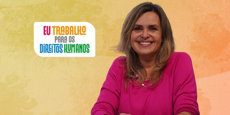 A imagem mostra uma mulher sorridente com cabelos loiros e ondulados, vestindo uma blusa rosa. Ela está sentada em frente a um fundo com tons de laranja e amarelo. À esquerda da mulher, há um texto colorido que diz: "Eu trabalho para os direitos humanos". As palavras "Eu trabalho" estão em laranja, "para os" em azul e "direitos humanos" em verde.