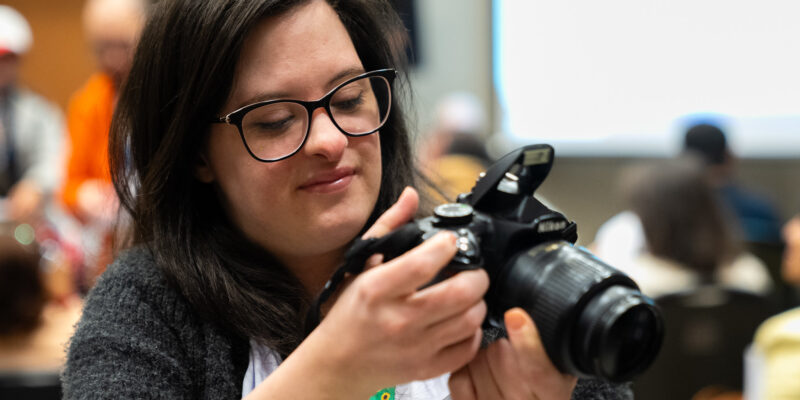 A imagem mostra uma mulher de cabelos castanhos escuros e longos, usando óculos de armação preta. Ela está segurando uma câmera fotográfica DSLR preta, da marca Nikon, e parece estar ajustando alguma configuração na câmera. A mulher está sorrindo levemente enquanto olha para a câmera. Ela veste uma blusa branca e um casaco cinza escuro. Ao fundo, há outras pessoas desfocadas, sugerindo que a cena ocorre em um ambiente interno, possivelmente durante um evento ou conferência.