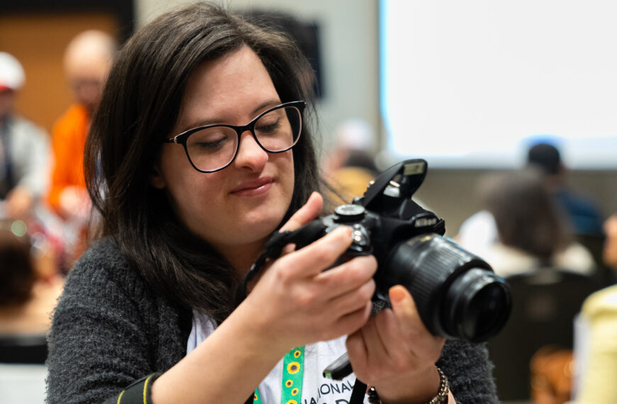 A imagem mostra uma mulher de cabelos castanhos escuros e longos, usando óculos de armação preta. Ela está segurando uma câmera fotográfica DSLR preta, da marca Nikon, e parece estar ajustando alguma configuração na câmera. A mulher está sorrindo levemente enquanto olha para a câmera. Ela veste uma blusa branca e um casaco cinza escuro. Ao fundo, há outras pessoas desfocadas, sugerindo que a cena ocorre em um ambiente interno, possivelmente durante um evento ou conferência.