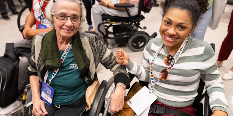 A imagem mostra duas mulheres sorrindo e sentadas em cadeiras de rodas. A mulher à esquerda é mais velha, tem cabelo grisalho e usa óculos. Ela está vestindo uma blusa verde com um casaco listrado em tons de cinza e verde, e um cachecol verde escuro. Ela tem um crachá pendurado no pescoço que diz "Porto Alegre". A mulher à direita é mais jovem, tem cabelo cacheado preso em um coque e está usando uma blusa de listras verdes e brancas. Ela também tem um crachá pendurado no pescoço e está segurando um par de óculos de sol. Ambas estão segurando as mãos e sorrindo para a câmera. Ao fundo, há outras pessoas, algumas também em cadeiras de rodas, em um ambiente que parece ser um local de evento ou conferência.