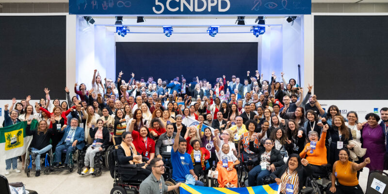 : A imagem mostra um grande grupo de pessoas reunidas em um evento, posando para uma foto. Elas estão em um ambiente interno, em frente a um palco com um letreiro que diz "5 CNDPD". Muitas pessoas estão sorrindo e levantando os braços em sinal de celebração. Há uma diversidade de participantes, incluindo pessoas em cadeiras de rodas. Algumas pessoas estão segurando bandeiras, uma delas é azul com detalhes amarelos, e outra parece ser a bandeira do estado do Rio Grande do Norte, que é verde com um brasão no centro. O ambiente parece ser um auditório ou centro de convenções, e há iluminação direcionada para o palco.