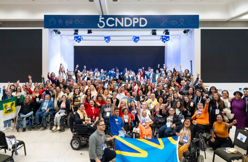 : A imagem mostra um grande grupo de pessoas reunidas em um evento, posando para uma foto. Elas estão em um ambiente interno, em frente a um palco com um letreiro que diz "5 CNDPD". Muitas pessoas estão sorrindo e levantando os braços em sinal de celebração. Há uma diversidade de participantes, incluindo pessoas em cadeiras de rodas. Algumas pessoas estão segurando bandeiras, uma delas é azul com detalhes amarelos, e outra parece ser a bandeira do estado do Rio Grande do Norte, que é verde com um brasão no centro. O ambiente parece ser um auditório ou centro de convenções, e há iluminação direcionada para o palco.