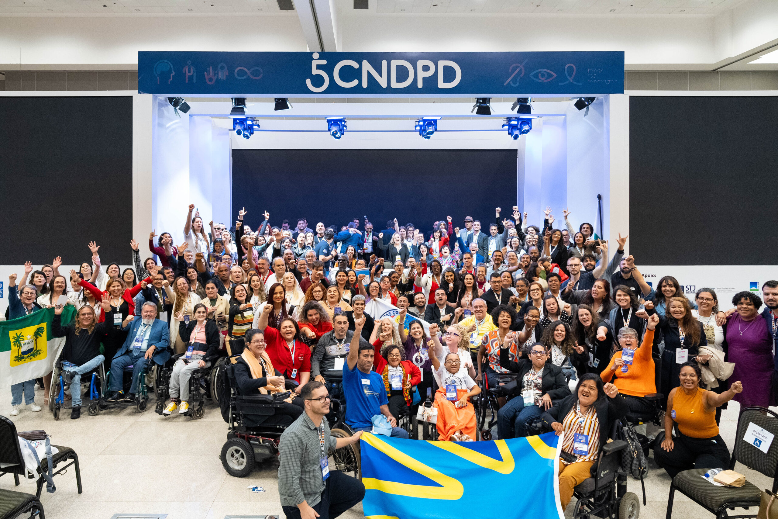 : A imagem mostra um grande grupo de pessoas reunidas em um evento, posando para uma foto. Elas estão em um ambiente interno, em frente a um palco com um letreiro que diz "5 CNDPD". Muitas pessoas estão sorrindo e levantando os braços em sinal de celebração. Há uma diversidade de participantes, incluindo pessoas em cadeiras de rodas. Algumas pessoas estão segurando bandeiras, uma delas é azul com detalhes amarelos, e outra parece ser a bandeira do estado do Rio Grande do Norte, que é verde com um brasão no centro. O ambiente parece ser um auditório ou centro de convenções, e há iluminação direcionada para o palco.