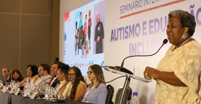 A imagem mostra um evento em um auditório, onde várias pessoas estão sentadas em uma mesa comprida, participando de um seminário. No canto direito da imagem, uma mulher negra está em pé, falando ao microfone. Ela veste uma roupa clara e parece estar fazendo uma apresentação ou discurso. Na parede atrás dela, há um grande banner com o título "Seminário Internacional Autismo e Educação Inclusiva". À esquerda, há uma projeção na parede com imagens de pessoas e um intérprete de língua de sinais. A mesa está coberta com uma toalha preta e há garrafas de água e microfones à frente dos participantes.