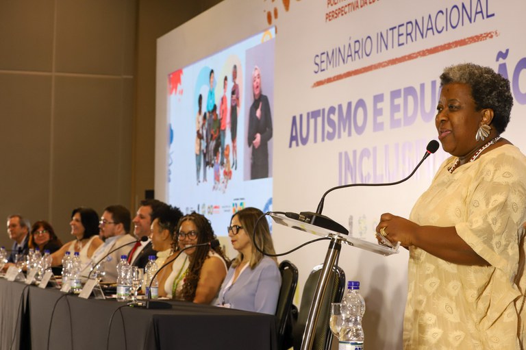 A imagem mostra um evento em um auditório, onde várias pessoas estão sentadas em uma mesa comprida, participando de um seminário. No canto direito da imagem, uma mulher negra está em pé, falando ao microfone. Ela veste uma roupa clara e parece estar fazendo uma apresentação ou discurso. Na parede atrás dela, há um grande banner com o título "Seminário Internacional Autismo e Educação Inclusiva". À esquerda, há uma projeção na parede com imagens de pessoas e um intérprete de língua de sinais. A mesa está coberta com uma toalha preta e há garrafas de água e microfones à frente dos participantes.