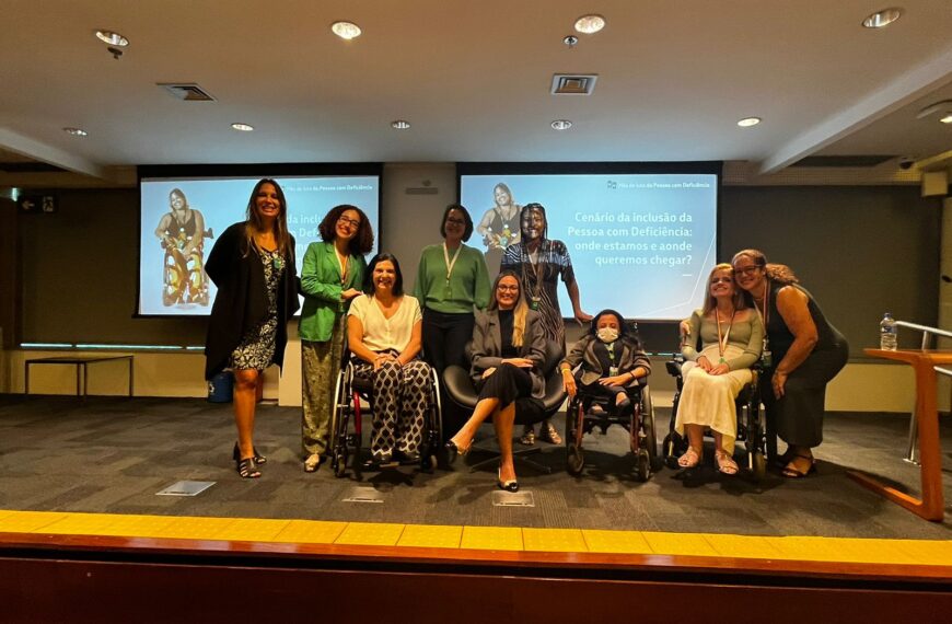 A imagem mostra um grupo de oito pessoas em um palco, posando para uma foto. Três delas estão em cadeiras de rodas. Ao fundo, há uma tela de projeção com a frase "Cenário da inclusão da Pessoa com Deficiência: onde estamos e aonde queremos chegar?". As pessoas estão sorrindo e parecem estar participando de um evento ou conferência sobre inclusão de pessoas com deficiência. O ambiente é uma sala de conferências com iluminação de teto.