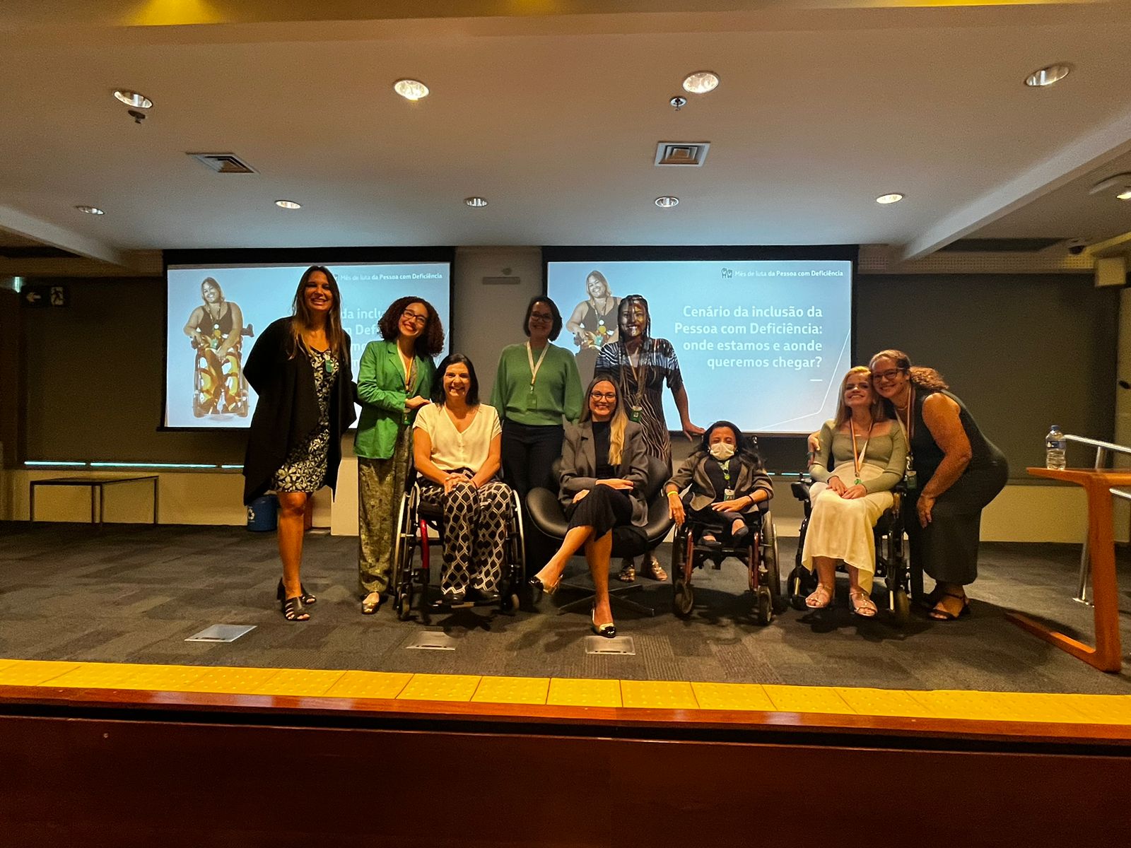 A imagem mostra um grupo de oito pessoas em um palco, posando para uma foto. Três delas estão em cadeiras de rodas. Ao fundo, há uma tela de projeção com a frase "Cenário da inclusão da Pessoa com Deficiência: onde estamos e aonde queremos chegar?". As pessoas estão sorrindo e parecem estar participando de um evento ou conferência sobre inclusão de pessoas com deficiência. O ambiente é uma sala de conferências com iluminação de teto.