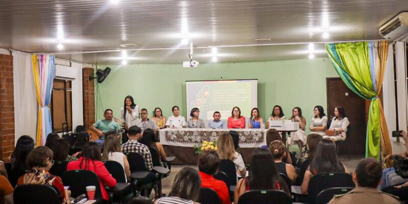 A imagem mostra uma sala de conferências com várias pessoas sentadas em cadeiras voltadas para um palco. No palco, há uma mesa comprida coberta com uma toalha branca e decorada com flores. Atrás da mesa, estão sentadas onze pessoas, a maioria mulheres, que parecem ser palestrantes ou membros de um painel. Uma das mulheres está falando ao microfone. Ao fundo, há uma tela de projeção com uma apresentação exibida. As paredes da sala são verdes e há cortinas coloridas nas laterais. A plateia está atenta, e algumas pessoas estão tirando fotos ou fazendo anotações. A iluminação é feita por lâmpadas no teto.