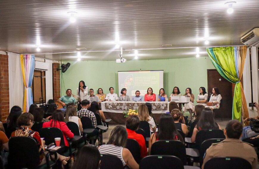 A imagem mostra uma sala de conferências com várias pessoas sentadas em cadeiras voltadas para um palco. No palco, há uma mesa comprida coberta com uma toalha branca e decorada com flores. Atrás da mesa, estão sentadas onze pessoas, a maioria mulheres, que parecem ser palestrantes ou membros de um painel. Uma das mulheres está falando ao microfone. Ao fundo, há uma tela de projeção com uma apresentação exibida. As paredes da sala são verdes e há cortinas coloridas nas laterais. A plateia está atenta, e algumas pessoas estão tirando fotos ou fazendo anotações. A iluminação é feita por lâmpadas no teto.