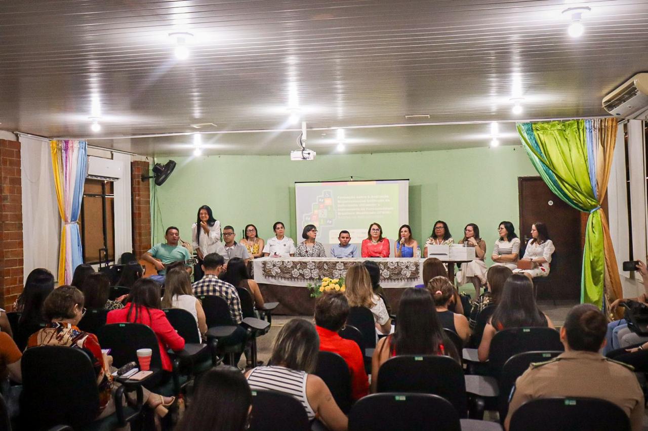 A imagem mostra uma sala de conferências com várias pessoas sentadas em cadeiras voltadas para um palco. No palco, há uma mesa comprida coberta com uma toalha branca e decorada com flores. Atrás da mesa, estão sentadas onze pessoas, a maioria mulheres, que parecem ser palestrantes ou membros de um painel. Uma das mulheres está falando ao microfone. Ao fundo, há uma tela de projeção com uma apresentação exibida. As paredes da sala são verdes e há cortinas coloridas nas laterais. A plateia está atenta, e algumas pessoas estão tirando fotos ou fazendo anotações. A iluminação é feita por lâmpadas no teto.