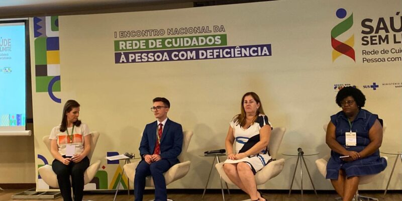 A imagem mostra quatro pessoas sentadas em cadeiras em um palco durante um evento. Da esquerda para a direita, a primeira pessoa é uma mulher de cabelo castanho, vestindo uma camiseta branca com um desenho colorido e calça preta. A segunda pessoa é um homem jovem, de óculos, vestindo um terno azul e gravata vermelha. A terceira pessoa é uma mulher de cabelo castanho claro, vestindo um vestido branco com detalhes em azul e preto. A quarta pessoa é uma mulher negra, de cabelo crespo, vestindo um vestido azul escuro. Todas as pessoas estão sentadas em cadeiras de cor clara, e há mesas pequenas ao lado de algumas delas com microfones. No fundo, há um banner com o texto "I Encontro Nacional da Rede de Cuidados à Pessoa com Deficiência". À direita, há o logotipo do evento "Saúde Sem Limite" e os logotipos do Brasil, SUS e Ministério da Saúde.