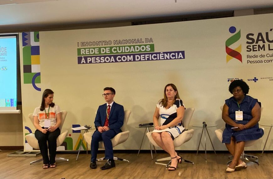 A imagem mostra quatro pessoas sentadas em cadeiras em um palco durante um evento. Da esquerda para a direita, a primeira pessoa é uma mulher de cabelo castanho, vestindo uma camiseta branca com um desenho colorido e calça preta. A segunda pessoa é um homem jovem, de óculos, vestindo um terno azul e gravata vermelha. A terceira pessoa é uma mulher de cabelo castanho claro, vestindo um vestido branco com detalhes em azul e preto. A quarta pessoa é uma mulher negra, de cabelo crespo, vestindo um vestido azul escuro. Todas as pessoas estão sentadas em cadeiras de cor clara, e há mesas pequenas ao lado de algumas delas com microfones. No fundo, há um banner com o texto "I Encontro Nacional da Rede de Cuidados à Pessoa com Deficiência". À direita, há o logotipo do evento "Saúde Sem Limite" e os logotipos do Brasil, SUS e Ministério da Saúde.