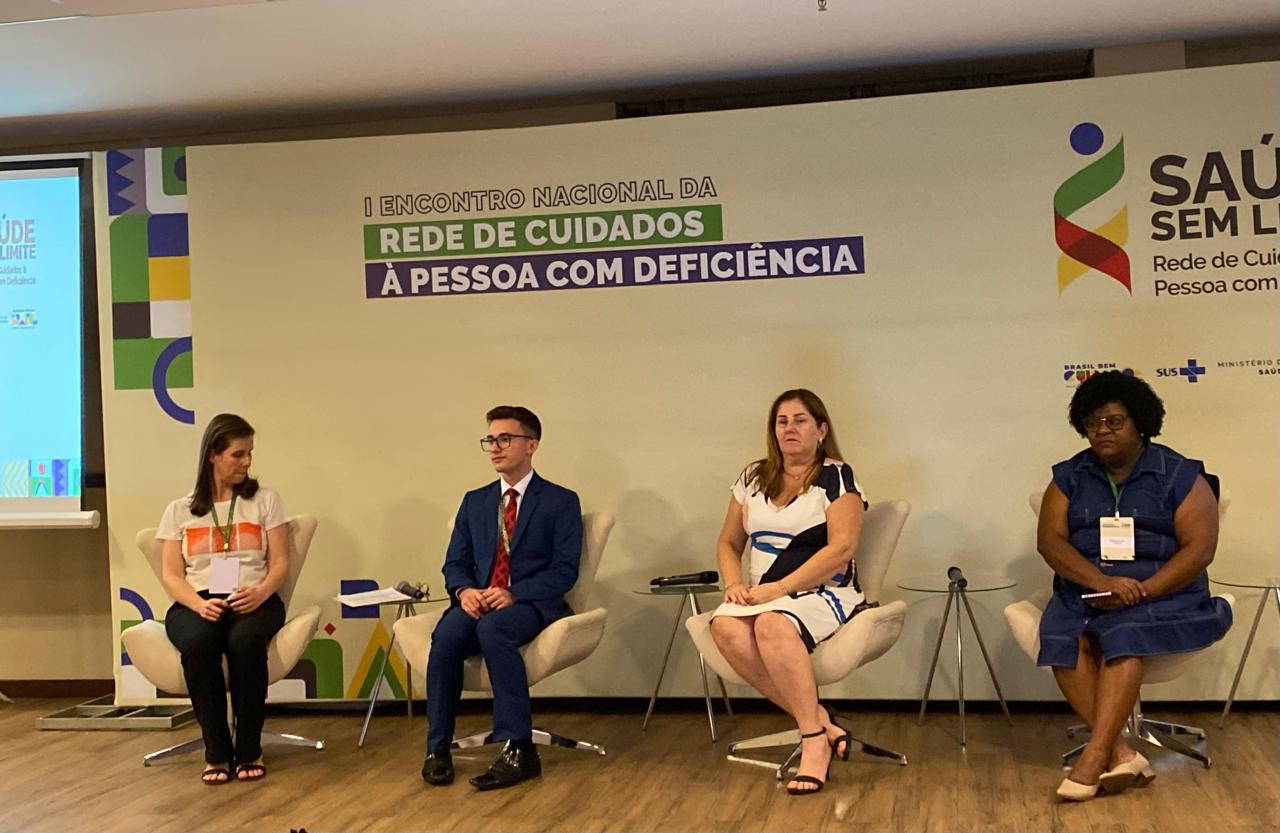 A imagem mostra quatro pessoas sentadas em cadeiras em um palco durante um evento. Da esquerda para a direita, a primeira pessoa é uma mulher de cabelo castanho, vestindo uma camiseta branca com um desenho colorido e calça preta. A segunda pessoa é um homem jovem, de óculos, vestindo um terno azul e gravata vermelha. A terceira pessoa é uma mulher de cabelo castanho claro, vestindo um vestido branco com detalhes em azul e preto. A quarta pessoa é uma mulher negra, de cabelo crespo, vestindo um vestido azul escuro. Todas as pessoas estão sentadas em cadeiras de cor clara, e há mesas pequenas ao lado de algumas delas com microfones. No fundo, há um banner com o texto "I Encontro Nacional da Rede de Cuidados à Pessoa com Deficiência". À direita, há o logotipo do evento "Saúde Sem Limite" e os logotipos do Brasil, SUS e Ministério da Saúde.